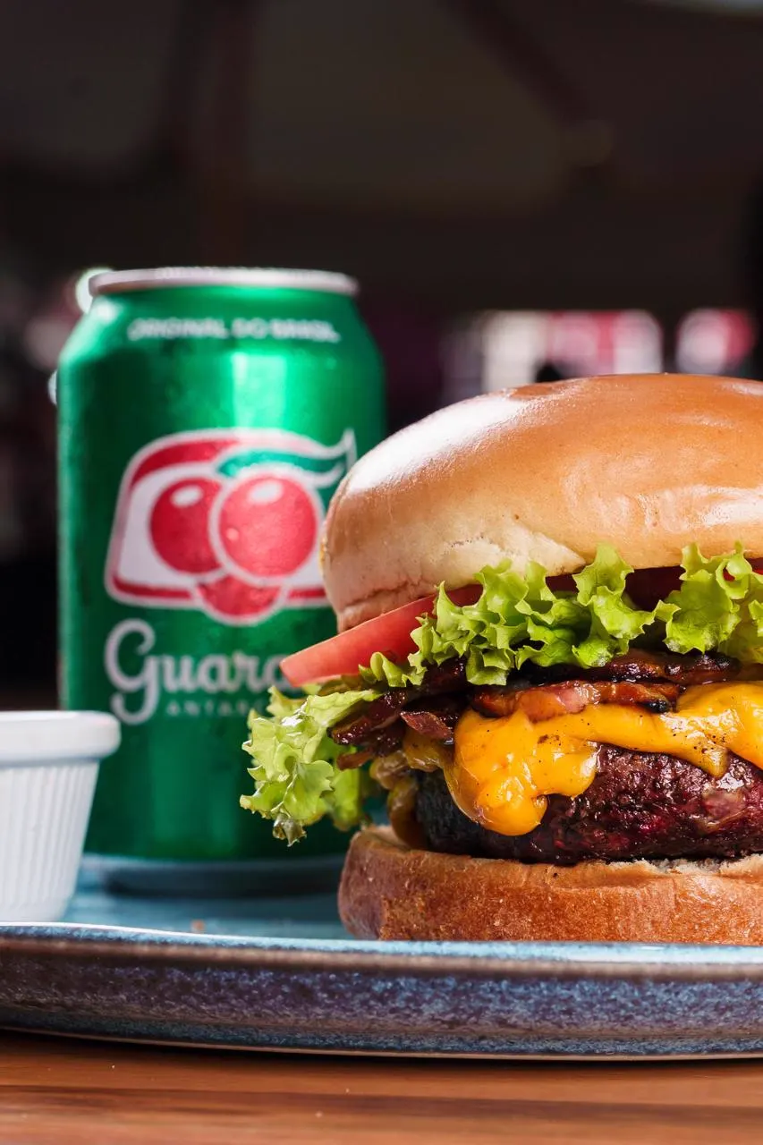 Hambúrguer com queijo cheddar, bacon, alface e tomate, com guaraná em lata ao fundo
