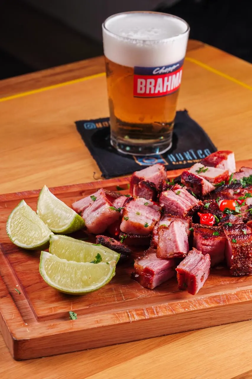 Torresmo em cubos na tábua com limão, atrás de um copo de chopp Brahma