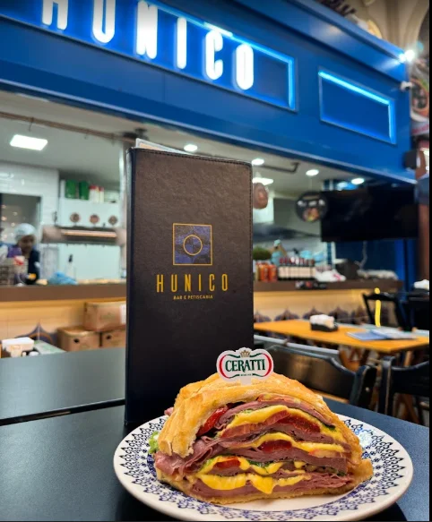 Lanche de mortadela com queijo diante da fachada azul do Hunico Mercadão